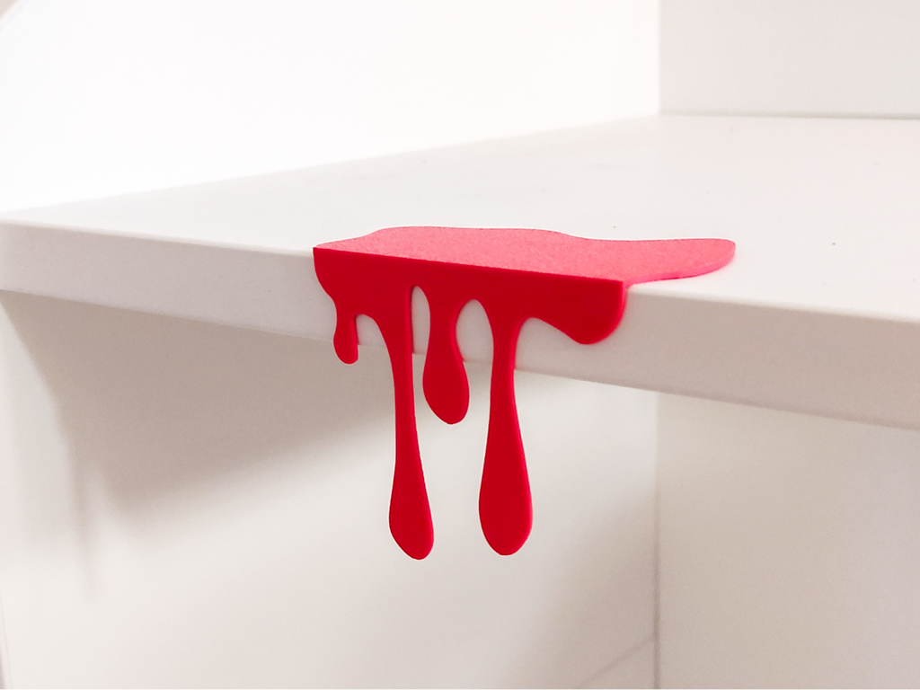 Red 3D printed blood drip decoration clipped onto white shelf edge, front view