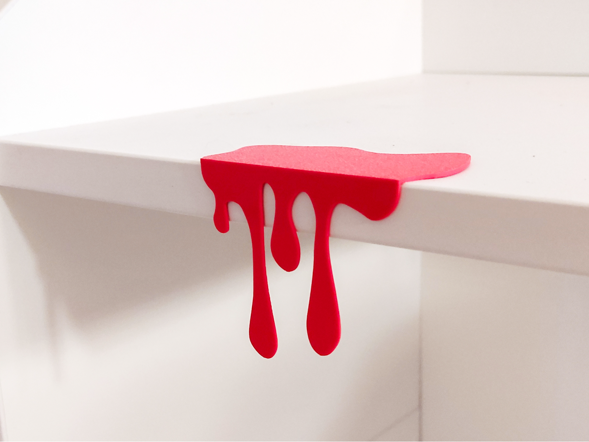 Red 3D printed blood drip decoration clipped onto white shelf edge, front view