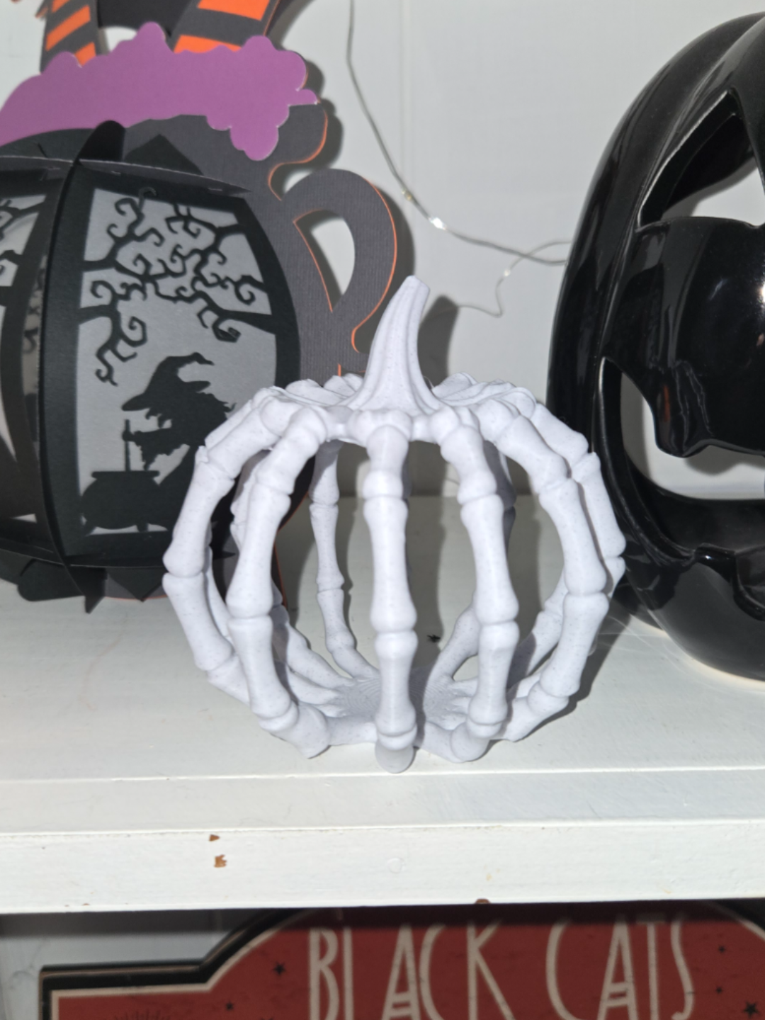 White 3D printed skeleton hand pumpkin on a white shelf surrounded by Halloween decor