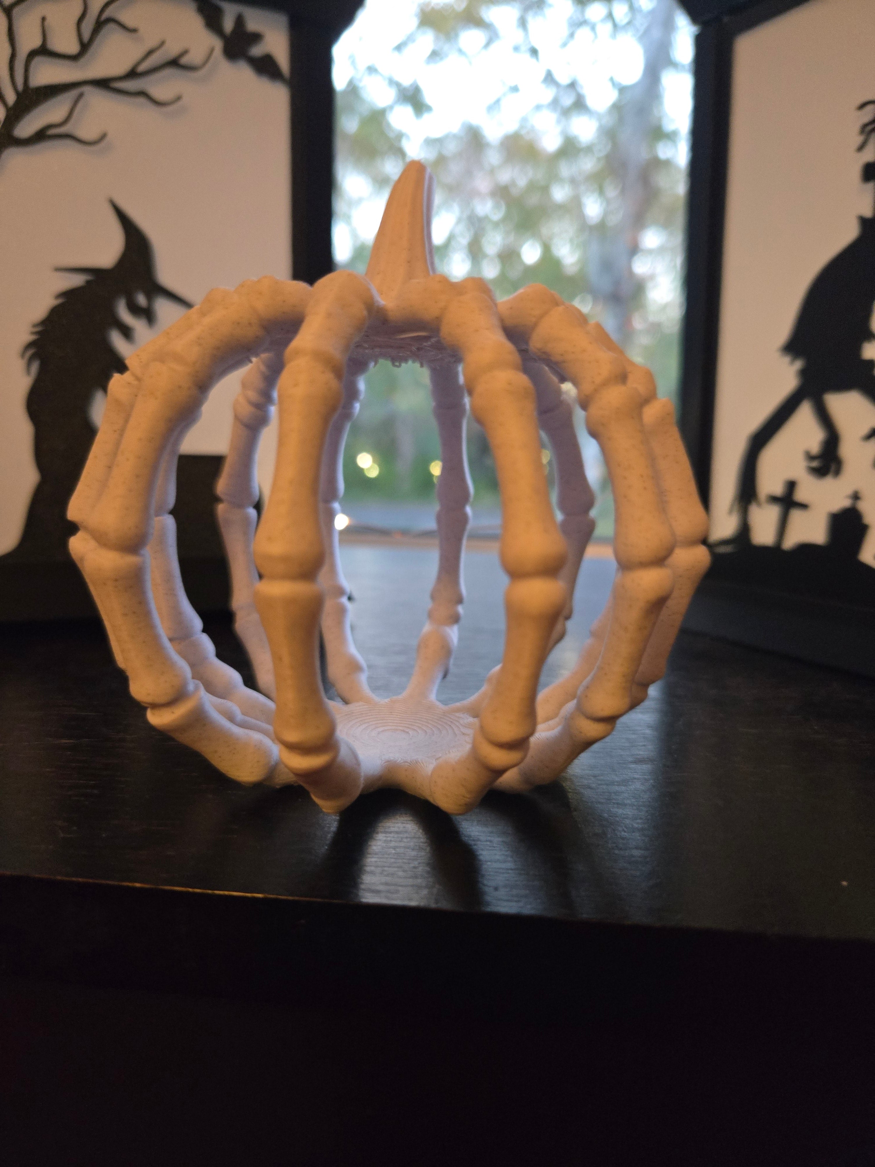 Cream 3D printed skeleton hand pumpkin on a dark surface with Halloween window silhouettes in background