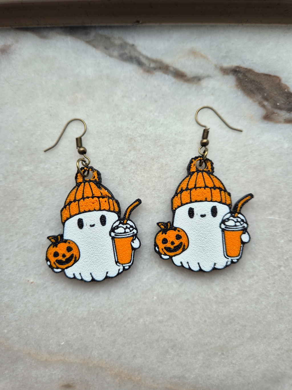 Pair of kawaii ghost Halloween earrings with orange beanie, pumpkin spice latte, and jack-o-lantern on marble surface