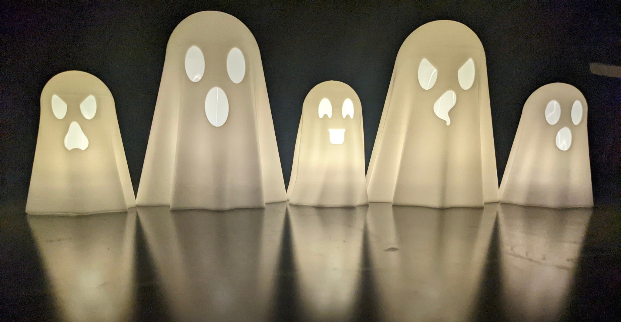 Five glowing 3D printed ghost lanterns with different face expressions lit up in a row on a reflective surface