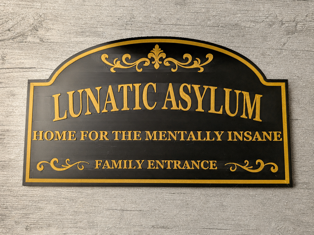 3D printed Lunatic Asylum Family Entrance novelty sign in black and gold on a grey wood wall
