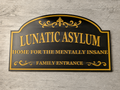 3D printed Lunatic Asylum Family Entrance novelty sign in black and gold on a grey wood wall