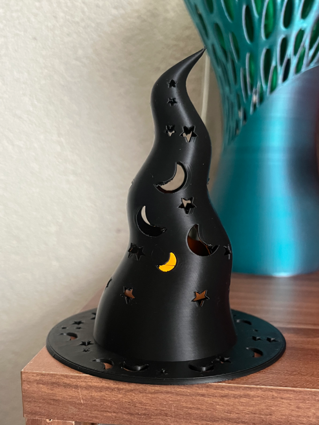Black 3D printed witch hat lantern with moon and star cutouts glowing warmly on wooden shelf
