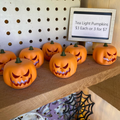 Multiple 3D printed orange jack-o-lantern tealight pumpkins glowing on a wooden shelf at a market display