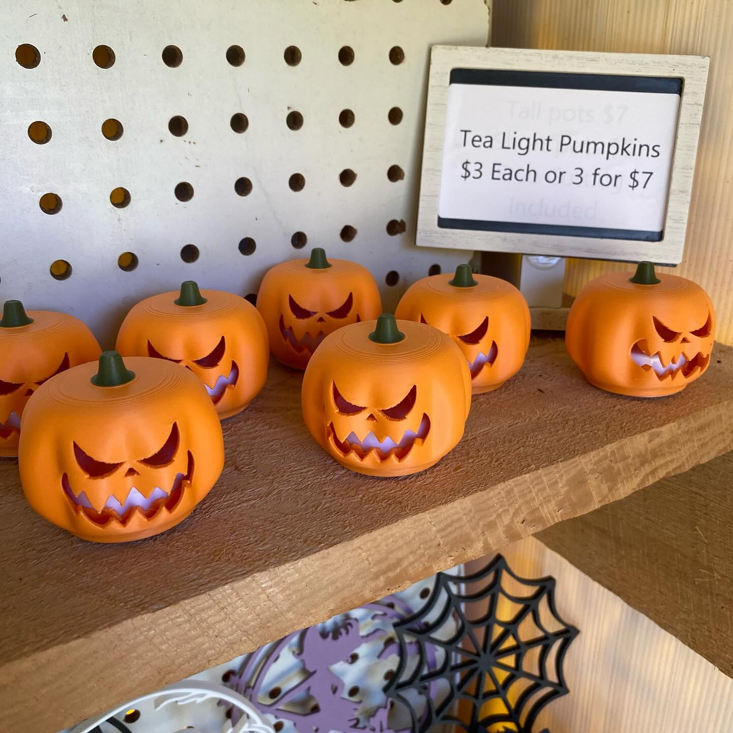 Multiple 3D printed orange jack-o-lantern tealight pumpkins glowing on a wooden shelf at a market display